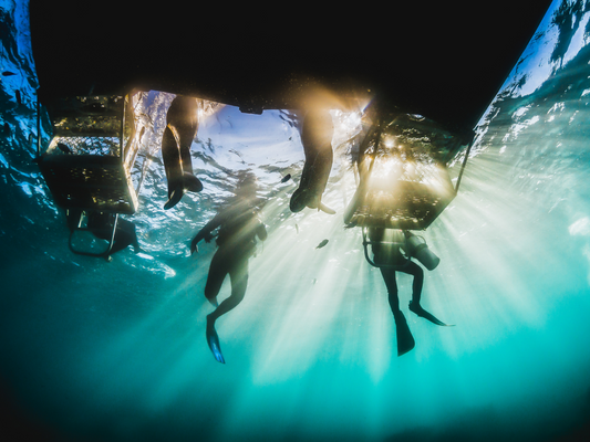 Underwater view of scuba divers swimming up to a boat as lights shine under the boat